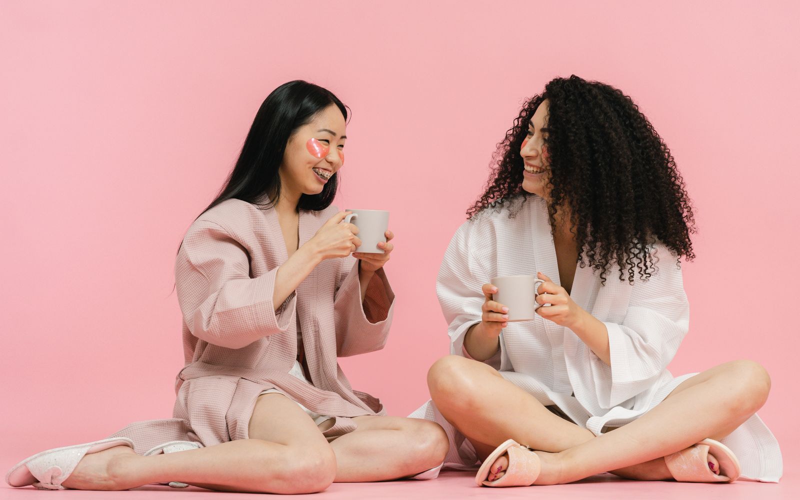 Two women in bathrobes relax with a warm drink, sitting on a pink background.