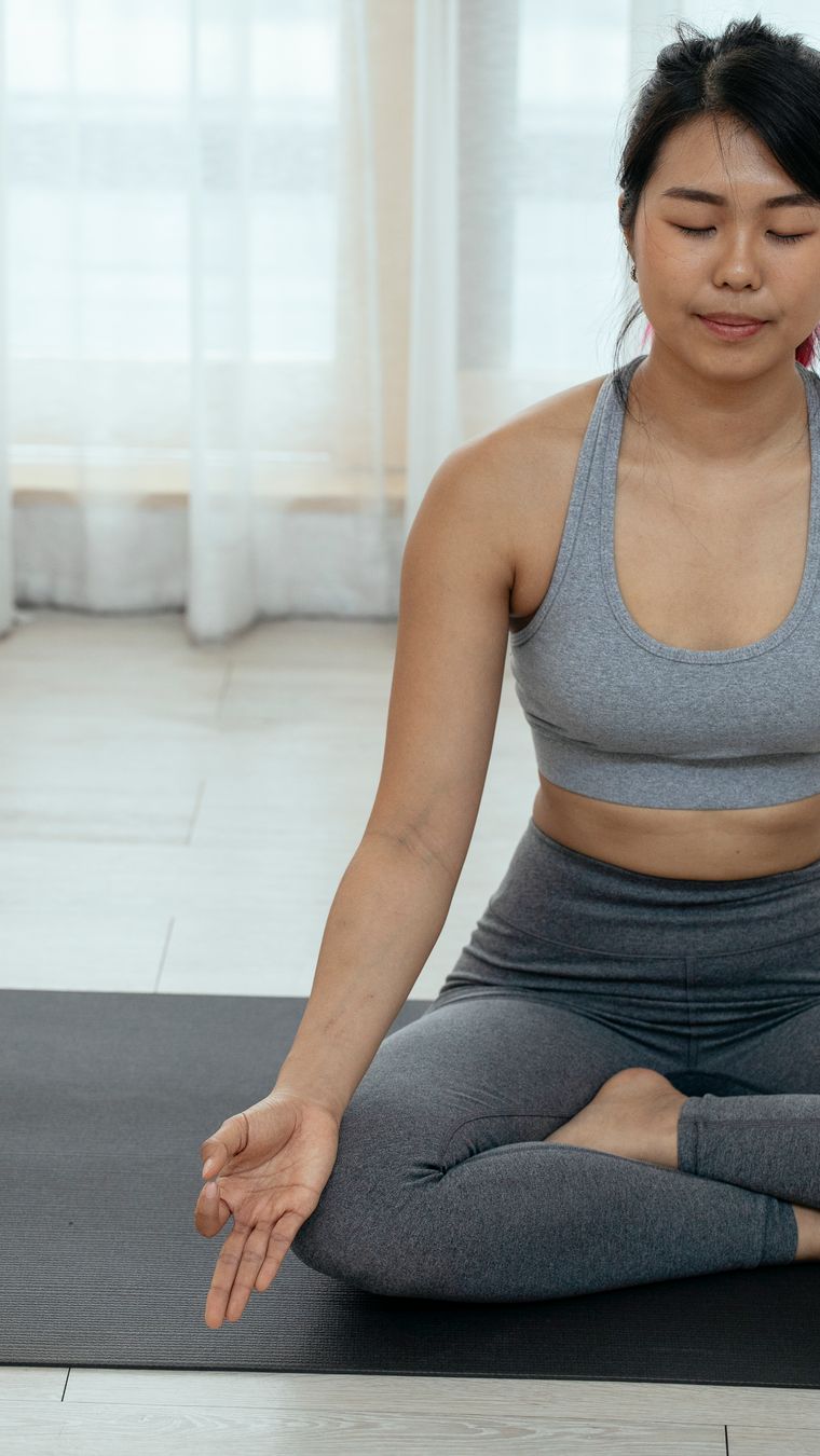 Asian woman in activewear sitting cross-legged, meditating on a yoga mat indoors.