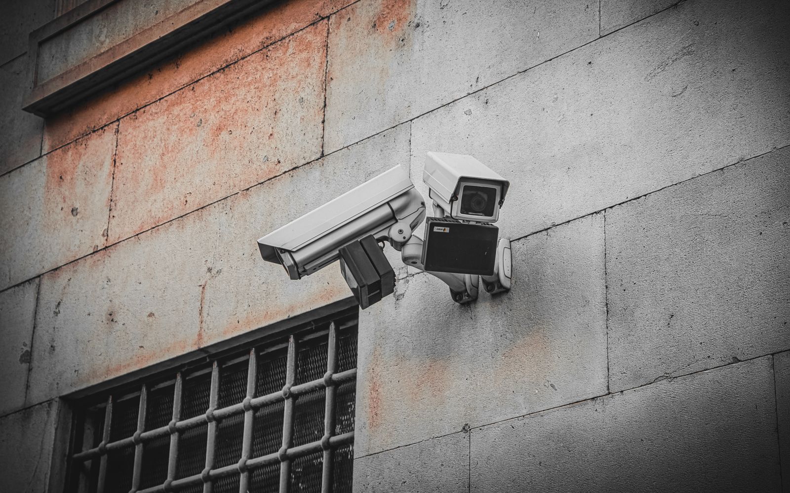 Two surveillance cameras mounted on an aged concrete wall beside a barred window, capturing urban security.