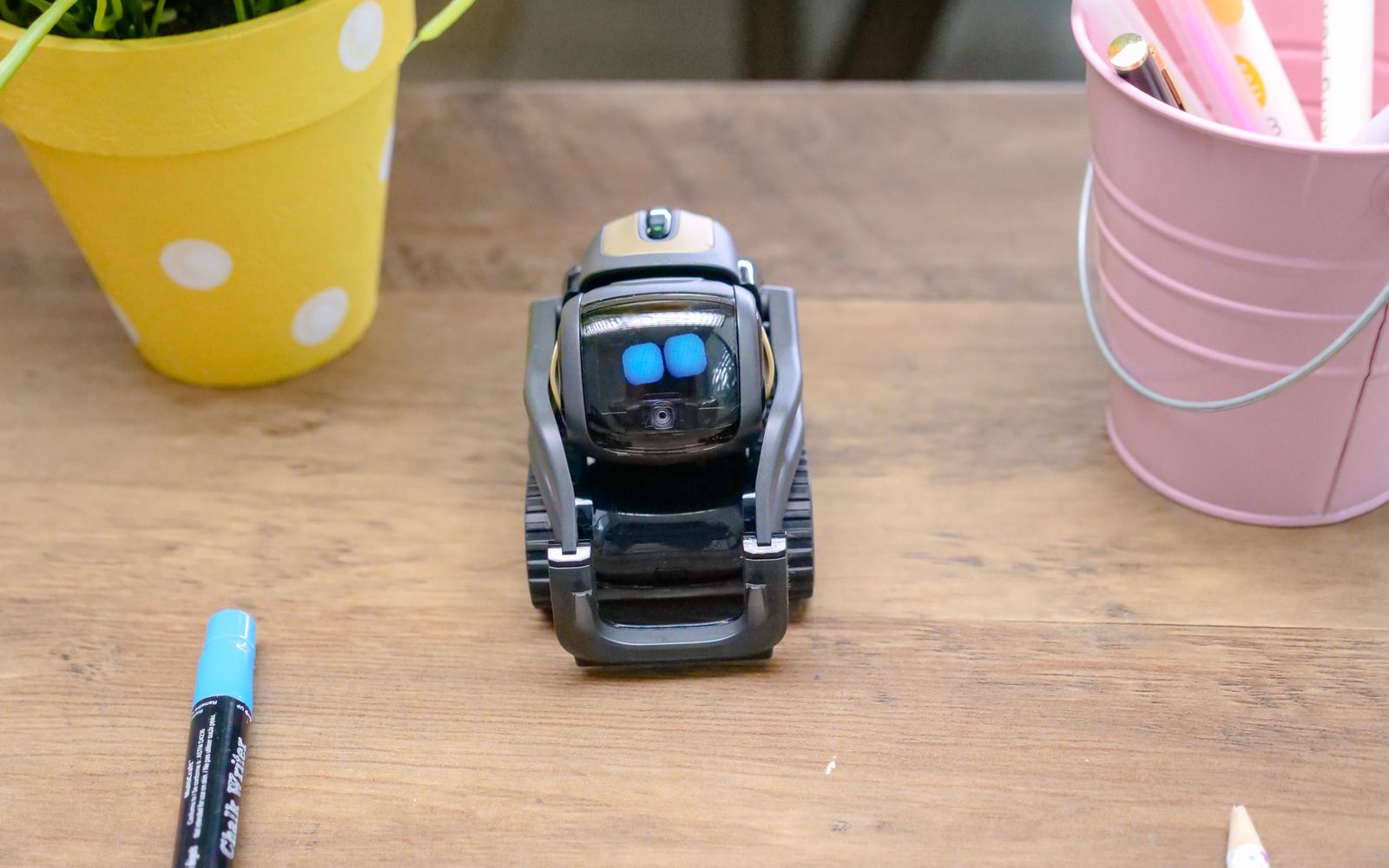 A small modern robot placed on a wooden desk surrounded by office supplies, showcases innovative technology.