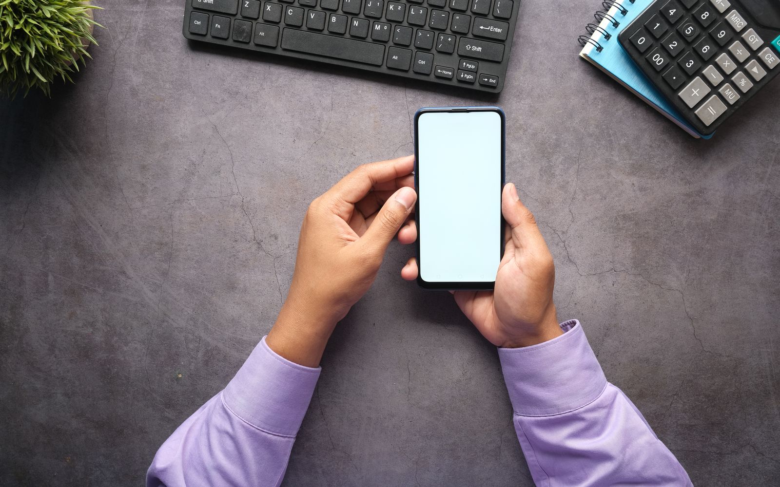 Man in purple shirt holding a smartphone with blank screen at office desk with keyboard, notebook, and calculator.